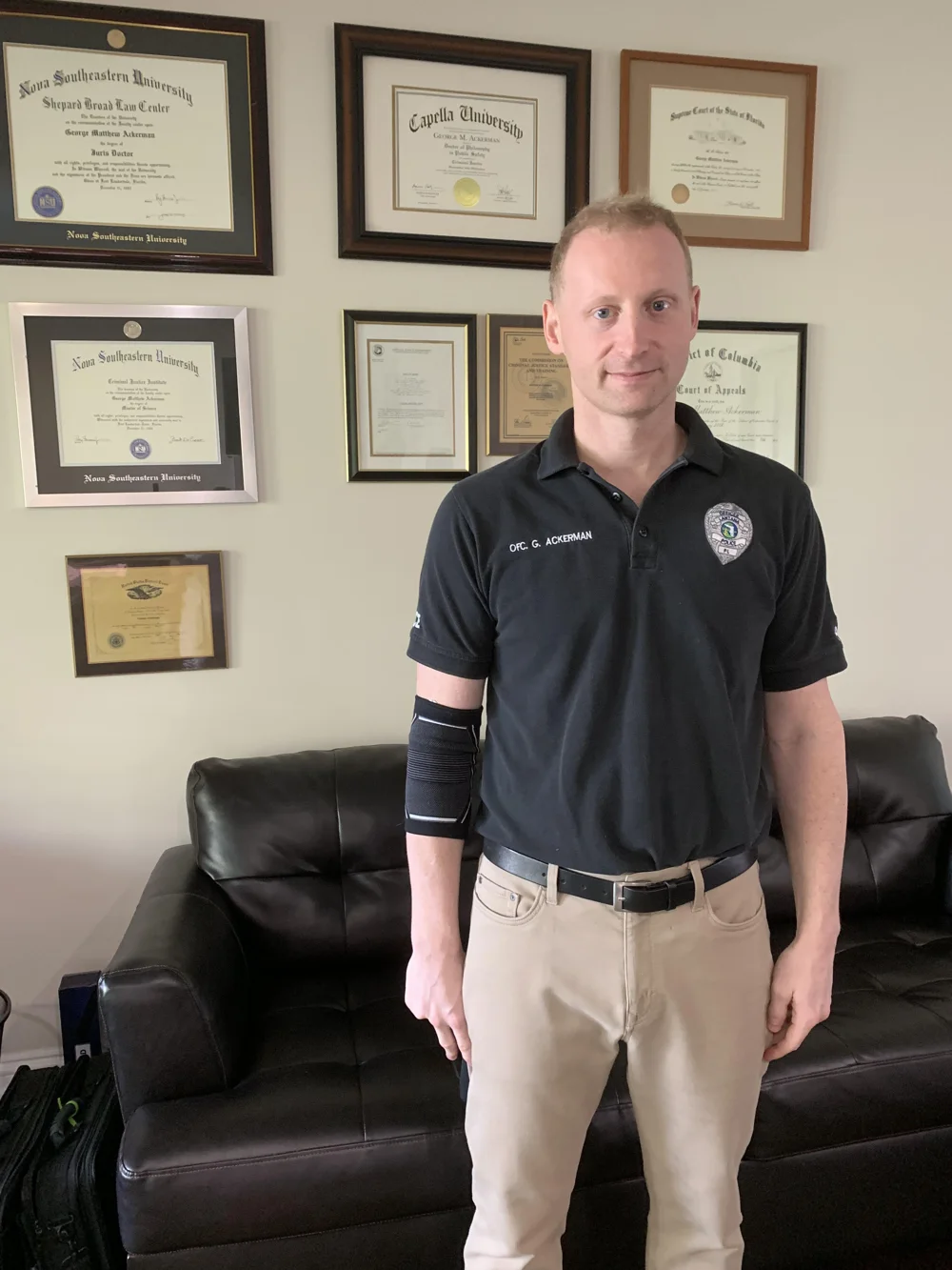 Dr. George Ackerman in a police department polo shirt in front of his framed degrees, 2019
